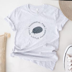 May include: A light gray t-shirt featuring a circular design with the text "THANKFUL FOR THE LITTLE THINGS" surrounding a gray pill bug illustration. The shirt is made of a soft material and has short sleeves.