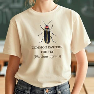 May include: Cream-colored t-shirt featuring a detailed illustration of a Common Eastern Firefly (Photinus pyralis). The firefly is depicted with a black body, yellow accents, and a red head. The text "COMMON EASTERN FIREFLY (Photinus pyralis)" is printed below the insect.