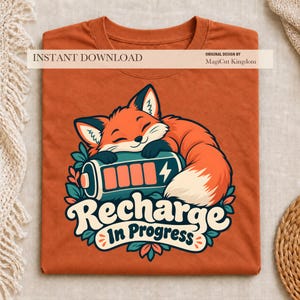 Può includere: Una t-shirt color arancio bruciato con la grafica di una volpe addormentata che riposa su un'icona a forma di batteria. Il testo sotto la volpe recita "Recharge In Progress" in un carattere color crema. Il design include foglie verde ottanio.