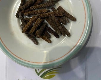 Puede incluir: Un cuenco de cerámica blanco con borde verde y rosa contiene una colección de especias secas, marrones y alargadas. Las especias están apiladas. Debajo del cuenco hay una tarjeta blanca con el texto "ALANG-ALANG Herb & Simplisia."