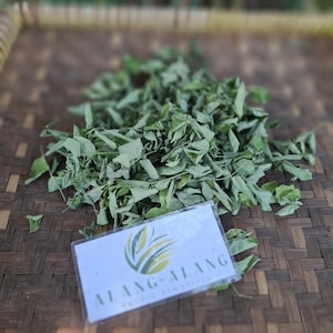 May include: A collection of dried green leaves, possibly herbs or tea, on a woven brown surface. A small white card with the text "ALANG-ALANG" and a green and yellow logo is in the foreground. The leaves are the main subject.