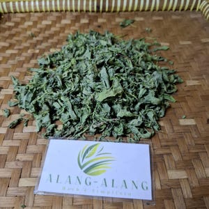 Könnte beinhalten: Getrocknete grüne Blätter auf einer geflochtenen Bambusoberfläche. Die Blätter liegen lose und verstreut. Ein weißes Etikett mit dem Text "ALANG-ALANG Herb & Simplisia" befindet sich in der unteren rechten Ecke des Bildes.