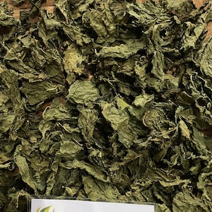 May include: A close-up of dried green leaves, possibly herbs, filling the frame. The leaves are textured and appear to be dried. A small, clear label at the bottom reads "Herbs & Simplista ALANG-ALANG" in white text with a green and yellow logo.