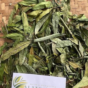 Puede incluir: Hojas verdes secas de alang-alang, un tipo de hierba, sobre una superficie de bambú tejido. Las hojas están dispuestas en una pila y una etiqueta blanca con el texto "Herb & Simplista ALANG-ALANG" es visible.