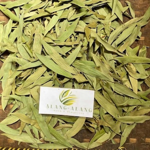 May include: A collection of dried, elongated, light green leaves spread out on a woven surface. A rectangular white label is placed on top of the leaves, featuring a green and yellow leaf logo and the text ALANG-ALANG Herb & Simplisia.