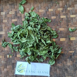 May include: A pile of dried green Alang-Alang leaves on a woven bamboo tray. A small white label with the text "ALANG-ALANG" is visible. The leaves have a crinkled texture and a natural appearance.