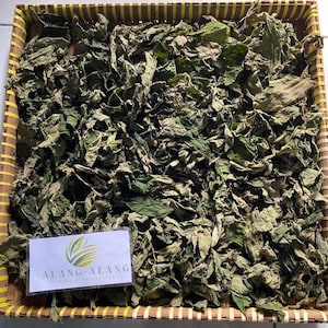 Puede incluir: Primer plano de una cesta tejida llena de hojas verdes secas. Las hojas están dispuestas en un patrón aleatorio y son de color verde claro. Una etiqueta blanca con el texto "ALANG-ALANG" es visible en la esquina inferior izquierda de la imagen.