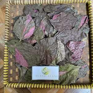May include: A collection of dried, textured leaves with variegated shades of dark brown, green, and magenta, arranged inside a square woven bamboo tray. A rectangular white label with a green leaf logo and the text ALANG-ALANG is placed on top.