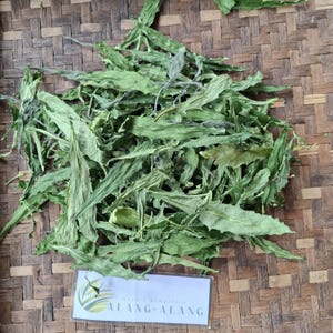 May include: A pile of dried green leaves, likely a type of herb, on a woven brown surface. A white label with the text "ALANG-ALANG" is visible in the lower right corner.