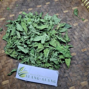 May include: A pile of dried green leaves, possibly herbs, on a woven bamboo surface. A label in the lower portion of the image reads "Herb & Simplisto ALANG-ALANG" with a green and yellow logo.