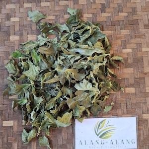May include: Dried green leaves, possibly herbs, are scattered on a woven surface. A small white card with the text "ALANG-ALANG Herb E Simplista" and a green logo is in the lower right corner. The leaves have a natural, organic appearance.