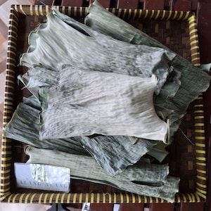 May include: A wicker tray contains several dried banana leaves, in shades of green and off-white. The leaves have a textured appearance with visible veins. A small white card with the text "ALANG-ALANG" is in the lower left corner.