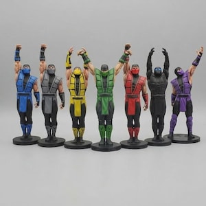 May include: A set of seven ninja figurines standing in a row on circular black bases. Each figure wears a sleeveless tunic, arm guards, and a face mask in a distinct color: blue, gray, yellow, green, red, black, and purple. All figures have both arms raised.
