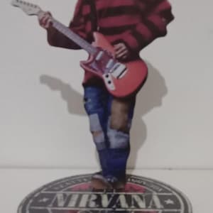 Puede incluir: Figura de un músico tocando una guitarra roja. La figura lleva una camisa a rayas rojas y negras y vaqueros azules. La base de la figura tiene el nombre de la banda "NIRVANA" y el año "1988".