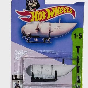 Könnte beinhalten: Ein Hot Wheels Spielzeugauto auf einer blau-grünen Karte. Das Auto ist weiß mit schwarzen Akzenten und dem Text "OceanGate". Die Karte zeigt das Hot Wheels-Logo, eine Darth Vader-Schädel-Grafik und den Text "TITAN DE OCEANGATE".