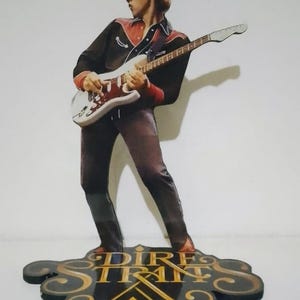 Puede incluir: Recorte de madera de un músico tocando una guitarra blanca. La figura lleva una camisa negra con detalles rojos y pantalones marrones. La base dice "Dire Straits" en letras doradas.