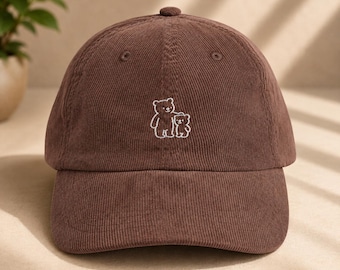 May include: A brown corduroy baseball cap featuring a small, white embroidered graphic of two bears standing side by side on the front panel. The cap has a structured crown, a curved brim, and a button at the top centre of the fabric panels.