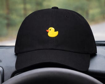 May include: Black baseball cap featuring a yellow rubber ducky emblem. The duck has an orange beak and is centrally placed on the cap. The cap is resting on a car dashboard, with a blurred backdrop of trees and a road.