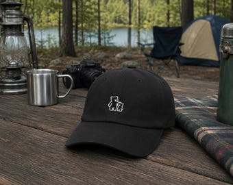 May include: A black baseball cap featuring a small, white, minimalist line-art graphic of a large bear standing next to a smaller bear. The cap has a curved brim and a structured crown, centred with the embroidered bear design on the front panel.