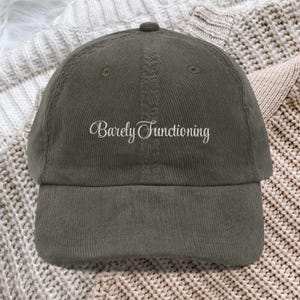 May include: A dark gray corduroy baseball cap featuring the embroidered white text Barely Functioning centered on the front panel. The cap has a structured crown, a curved brim, and a button at the top, showcasing a textured fabric finish.