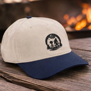 May include: A beige baseball cap with a dark blue brim and a dark blue button on top. The front features a black circular graphic depicting a silhouette of an alien and Bigfoot walking together under a flying saucer with stars and pine trees.