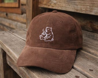 May include: Brown corduroy baseball cap with a white embroidered bear drinking from a cup. The cap has a curved peak and a classic six-panel design. The hat is resting on a wooden bench.