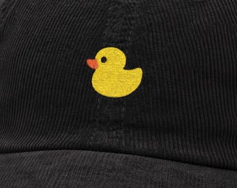 May include: Black corduroy baseball cap with a yellow duck design and an orange beak. The cap has a curved peak and a fabric-covered button. The duck is embroidered on the front.