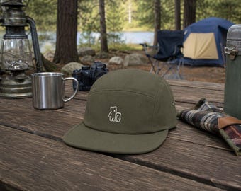 May include: An olive green five-panel cap featuring a small, white, minimalist embroidered graphic of a large bear standing next to a smaller bear on the front centre panel. The hat has a flat brim and a structured crown with a side metal eyelet.