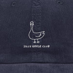 May include: Navy blue corduroy baseball cap featuring a white embroidered goose design and the text "SILLY GOOSE CLUB". The cap has a curved brim and a structured crown. A casual accessory.
