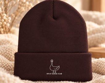 May include: A dark brown knit beanie featuring a white embroidered graphic of a goose and the text "SILLY GOOSE CLUB" centred on the folded cuff. The hat has a ribbed texture and a classic cuffed design, providing a simple and clean look.