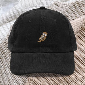 May include: A black corduroy baseball cap featuring an embroidered design of a brown otter holding a smaller otter on its chest. The cap has a structured crown, a curved brim, and a button on top, showcasing a textured fabric finish throughout.