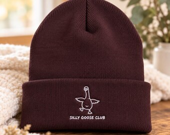 May include: A dark brown knit beanie featuring a white embroidered graphic of a goose with its wings spread and the text "SILLY GOOSE CLUB" centred on the folded cuff. The hat is made of a ribbed material with a classic cuffed design.