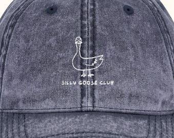 May include: A denim blue baseball cap with a white outline of a goose wearing a hat. The text "SILLY GOOSE CLUB" is below the goose. The cap has a curved peak and a dark blue button on top.