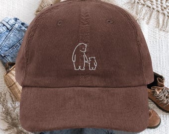 May include: A brown corduroy baseball cap featuring a white embroidered line art graphic of a large bear holding the hand of a small bear. The cap has a structured crown, a curved brim, and a button on top, showcasing a textured fabric finish.