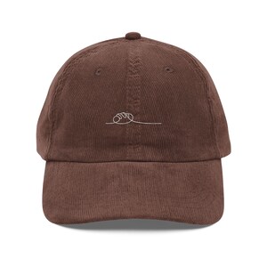 May include: A brown corduroy baseball cap with a curved peak. The cap has a small, white, minimalist line drawing of a loaf of bread. The cap has a six-panel design with a top button.