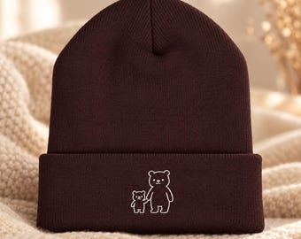 May include: A dark brown knit beanie featuring a white embroidered graphic of a large bear holding the hand of a small bear. The hat has a folded cuff design and a ribbed texture throughout the fabric, showcasing a simple line art illustration.