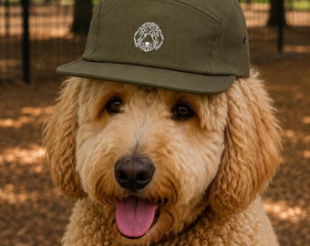 May include: A golden-coloured dog wearing an olive green five-panel cap with a white embroidered dog face. The dog has a pink tongue and a bone-shaped tag on its collar. The background is a park with trees.