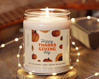 May include: A lit candle in a glass jar with a label that reads "Happy Thanksgiving Day." The label features illustrations of pumpkins and sunflowers. The candle sits on a wooden coaster and is surrounded by string lights.