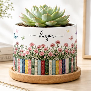 Op de afbeelding: Een witte cilindrische keramische plantenbak met de naam Harper in zwarte schrijfletters, versierd met een patroon van roze anjers, kleurrijke vlinders en een rij verticale boekruggen aan de onderkant, op een ronde houten schotel.