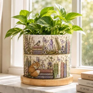 Pode incluir: Um vaso cilíndrico de cerâmica com fundo creme, apresentando uma ilustração detalhada de uma raposa, livros empilhados, borboletas e cristais. O vaso contém uma planta verde e repousa sobre uma base circular de madeira clara.