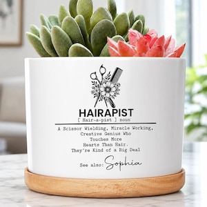 May include: A white ceramic planter with a wooden base containing green and pink succulents. The planter features a black graphic of scissors and a comb with the text: HAIRAPIST, A Scissor Wielding, Miracle Working, Creative Genius Who Touches More Hearts Than Hair. They're Kind of a Big Deal. See also: Sophia.