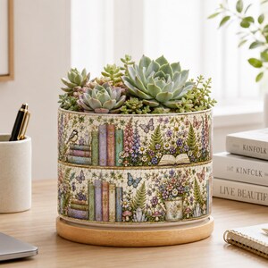 Pode incluir: Um vaso cilíndrico de cerâmica com uma ilustração detalhada de livros empilhados, flores silvestres, borboletas e um regador. O vaso contém várias suculentas verdes e assenta sobre uma base circular de madeira clara.