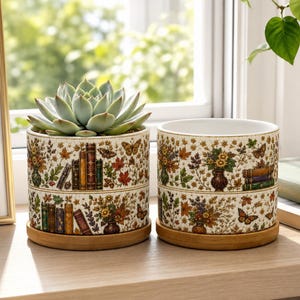 Può includere: Due vasi cilindrici in ceramica con sottovasi in bambù, caratterizzati da uno sfondo color crema con un motivo ripetuto di libri vintage colorati, foglie autunnali, composizioni floreali e farfalle. Un vaso contiene una pianta succulenta.