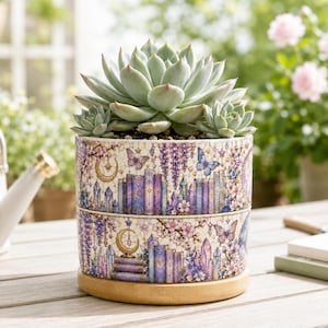 Op de afbeelding: Een cilindervormige keramische plantenpot met een groene vetplant, voorzien van een decoratieve print van paarse boeken, kristallen, vlinders, maansikkels en bloemenranken. De pot staat op een ronde, lichtgekleurde houten basis.
