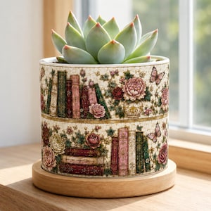 May include: A cylindrical ceramic planter featuring a detailed print of stacked books, blooming flowers, and butterflies in shades of pink, green, and gold. A light green succulent plant sits inside the pot, which rests on a circular wooden base.