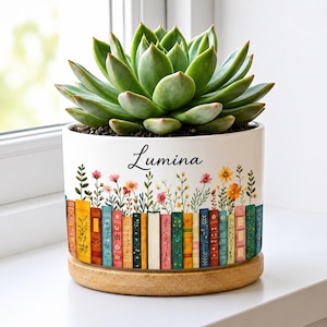 May include: A white ceramic cylindrical planter containing a green succulent. The planter features a colorful illustration of a row of books with wildflowers growing from the top and the name Lumina printed in black script. It sits on a wooden base.