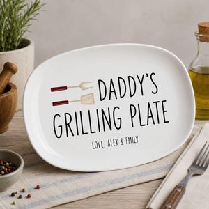 Peut inclure: Plat ovale blanc avec l'inscription noire "DADDY'S GRILLING PLATE". Le design comprend une fourchette et une spatule avec des manches rouges. Le bas de l'assiette indique "LOVE, ALEX & EMILY".
