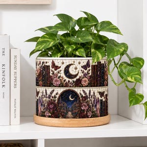 May include: A cylindrical ceramic planter featuring a detailed wrap-around illustration of a moonlit garden with roses, butterflies, and books. The planter sits on a circular wooden base and contains a trailing green pothos plant with variegated leaves.