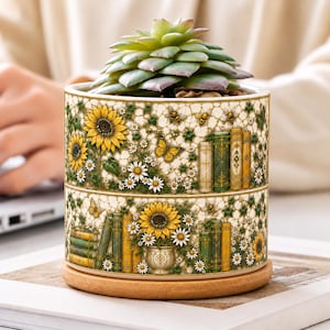 Può includere: Un vaso cilindrico in ceramica con un motivo dettagliato di girasoli gialli, margherite bianche, libri antichi verdi e dorati, piccole api e farfalle. Una pianta succulenta verde è posta all'interno del vaso su una base in legno.