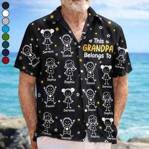 Op de afbeelding: Zwart overhemd met een patroon van stokfiguren, sterren en namen. De tekst "This Grandpa Belongs To" is in goud gedrukt. Het shirt heeft korte mouwen en een klassieke kraag.